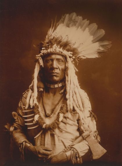 Cua de Mostela, guerrer dels indis piegan, retratat per Curtis el 1900. Porta un barret de plomes d'àliga i un collaret d'urpes d'ós grizzly, i va armat amb un tomahawk.